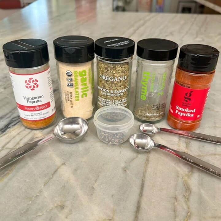 Row of five spice jars including two kinds of paprika on a marble countertop with metal measuring spoons.