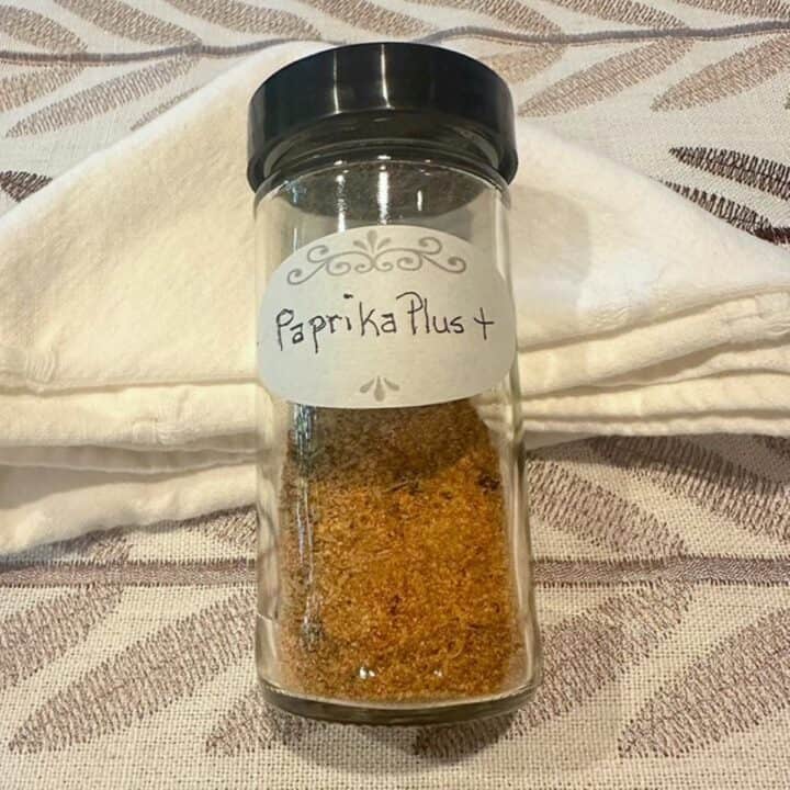 A clear spice jar with reddish dried spices lays on a beige, leaf-patterned tablecloth.