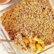 A golden apple crisp fills a clear, square baking dish with a serving spoon digging in.