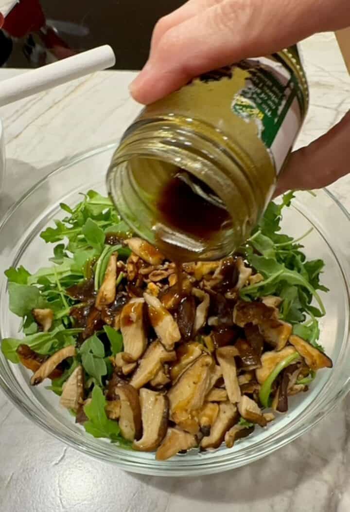 Pouring dark brown dressing over arugula, mushrooms, and walnuts in a clear bowl.