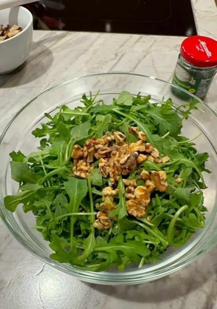 A clear bowl holds a mound of leafy green arugula topped with brown, toasted walnuts. 