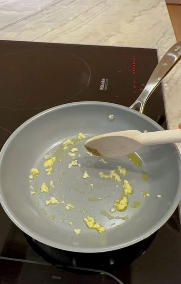 Golden minced garlic is cooked in a gray non-stick pan in hot oil on a black stovetop. 