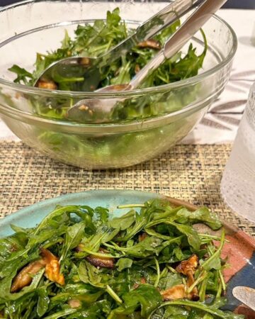 Foreground with blue-green plate holding a tangle of green arugula with walnuts and mushrooms. Clear bow with the arugula salad in the background.