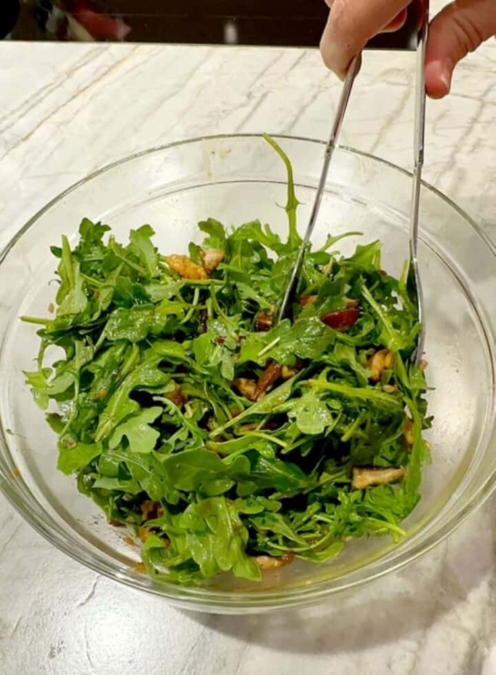 Green arugula leaves with sliced, cooked brown mushrooms in a clear bowl with silver tongs.
