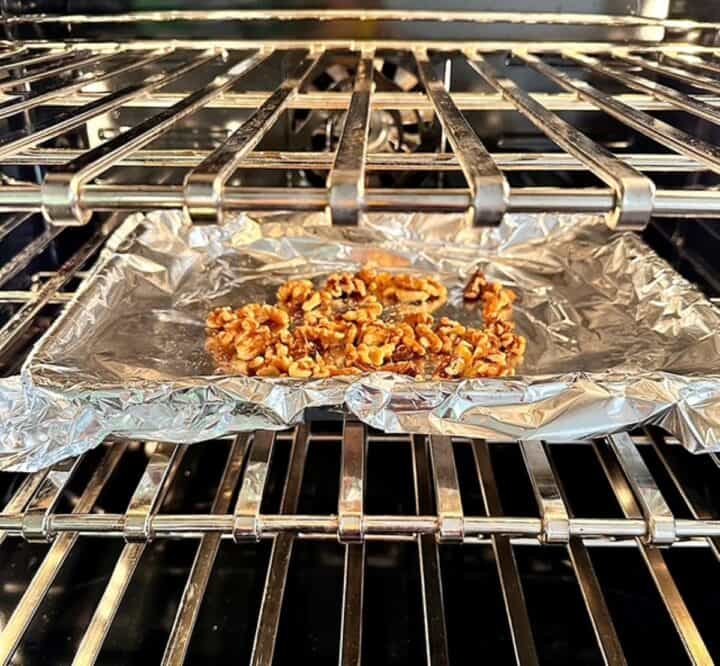 Looking inside an oven, a shiny, foil-covered sheet pan holds brown walnuts, toasting.