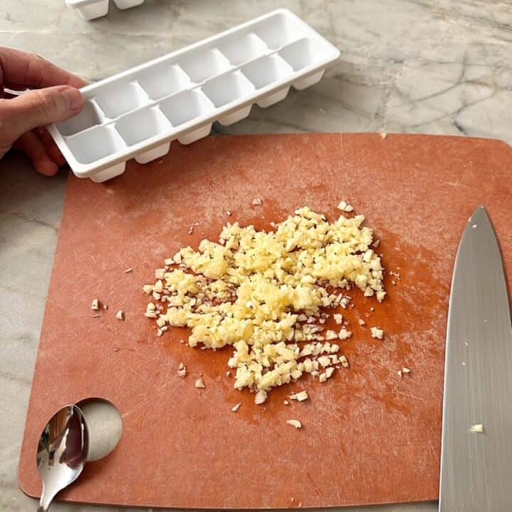 Brown cutting board with a large amount of creamy colored chopped garlic, an empty white ice cube tray on the side.