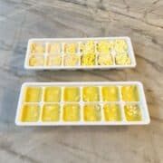 Two small white ice cube trays with frozen cubes of dark yellow grated ginger and a lighter yellow chopped garlic.