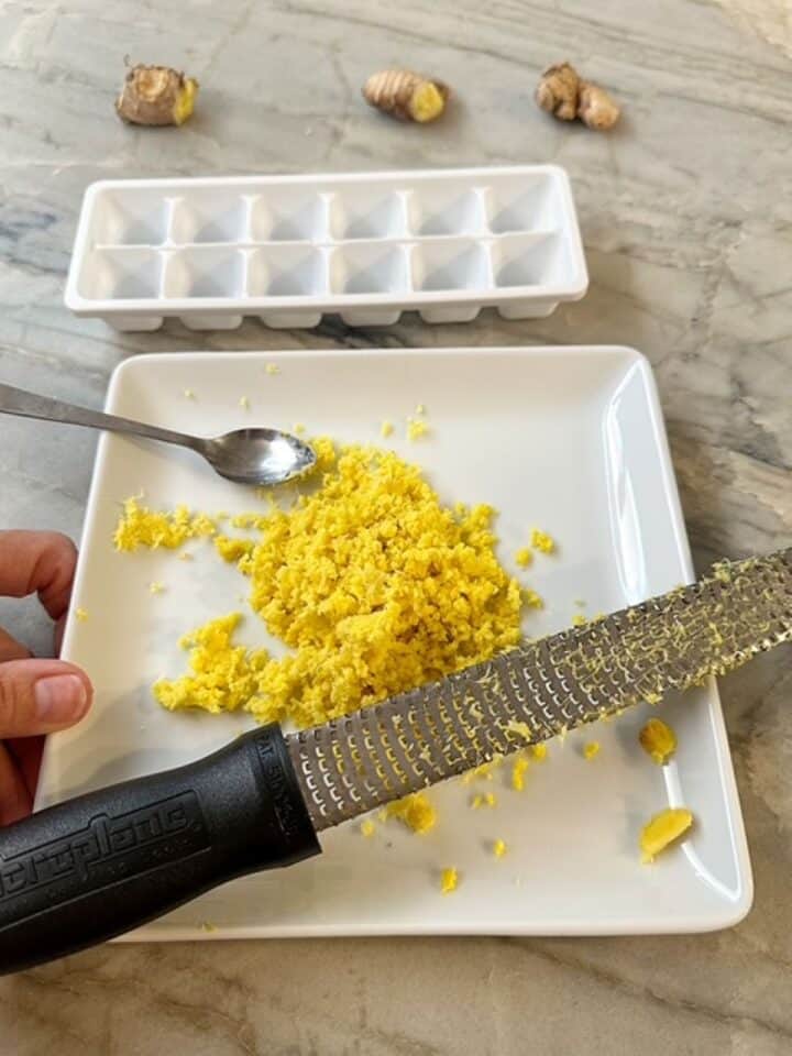 A square white plate holds a heap of grated ginger, a small spoon, and a microplane grater
