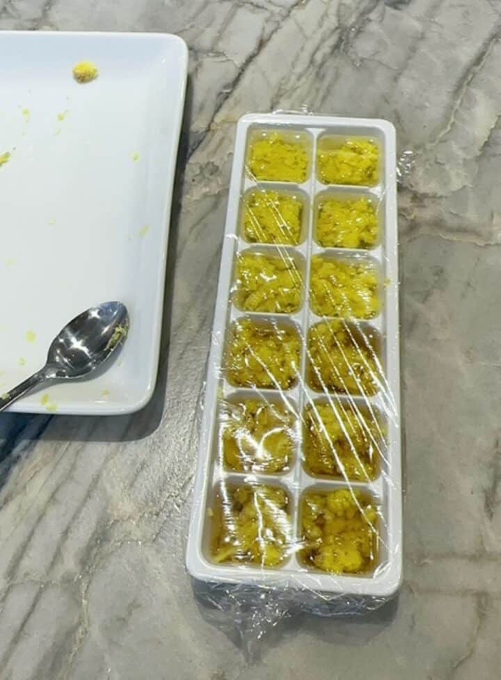Small white ice cube tray filled with yellow grated ginger in oil, covered with clear plastic wrap.