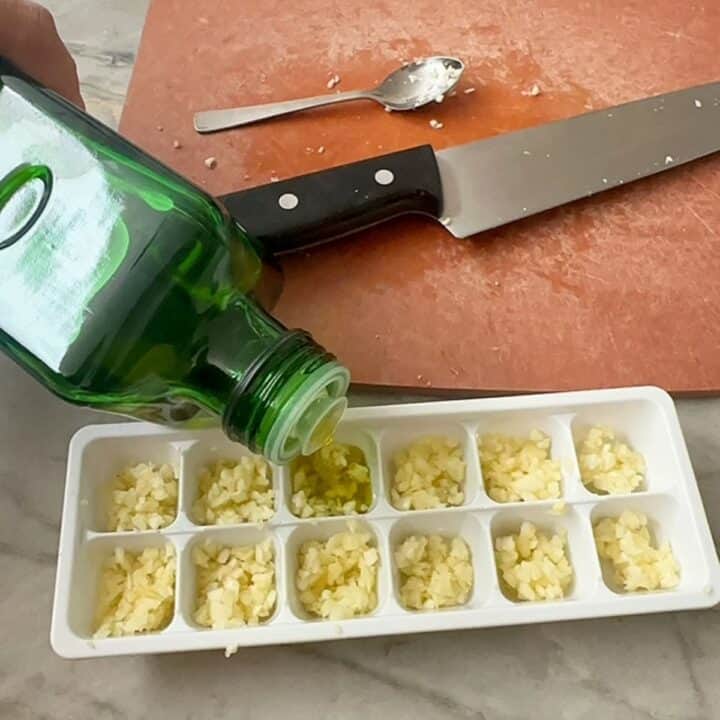 Pouring from green olive oil bottle to cover yellowish chopped garlic in a white ice cube tray.