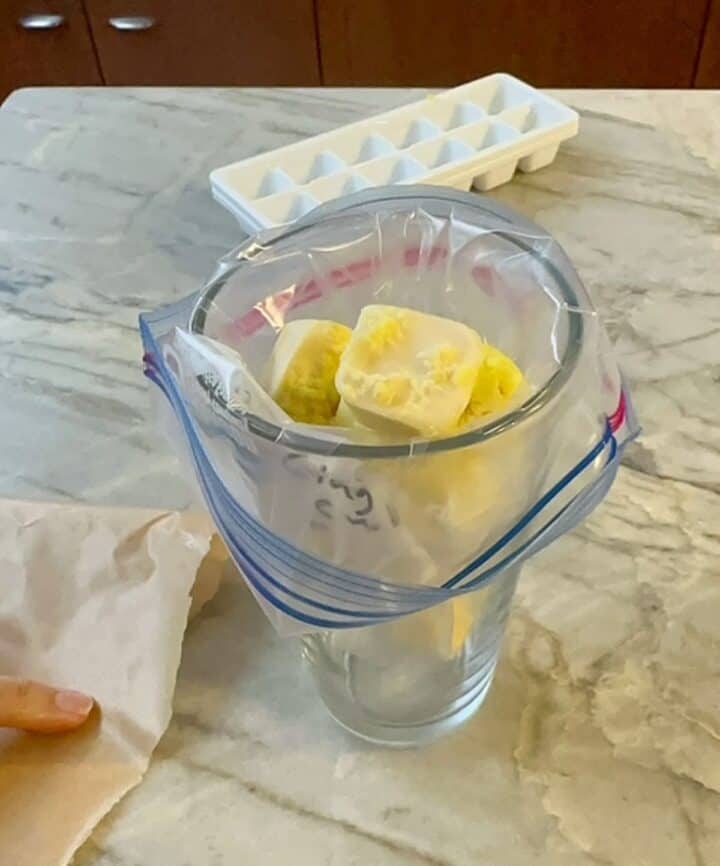 A zip bag full of yellow frozen garlic cubes is perched in tall, clear glass.