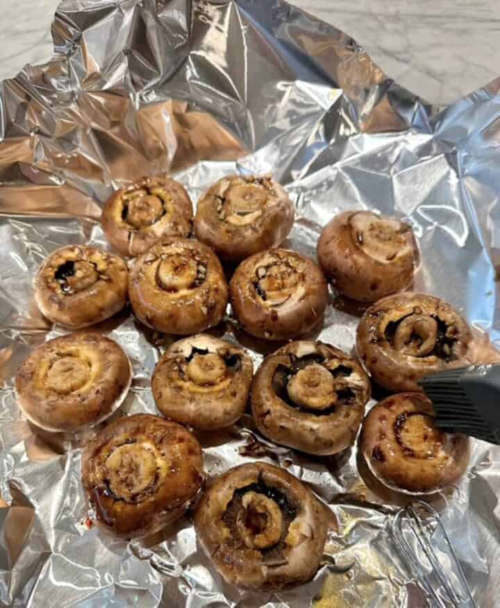 Brown cremini mushrooms are crowded together, cap side down, on a piece of shiny aluminum foil.