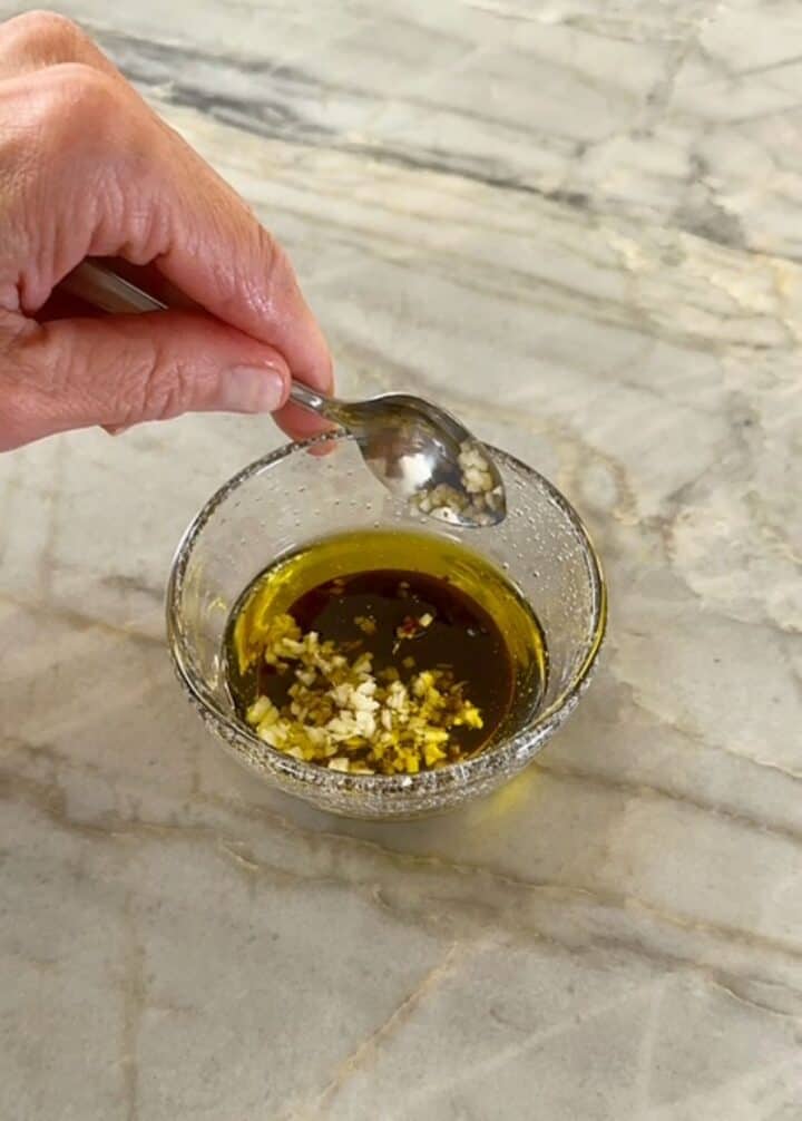 Hand holds a spoon over a mall, clear bowl with green olive oil, brown balsamic vinegar, and creamy garlic.