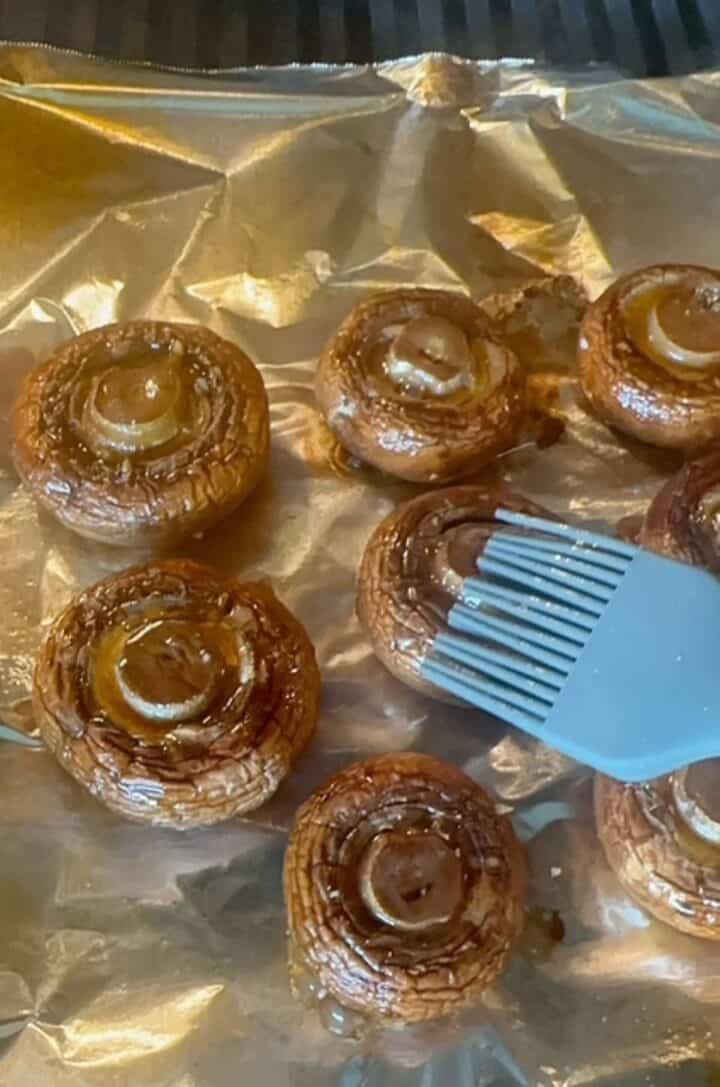 A blue silicone pastry brush coats brown creminis on foil with balsamic glaze.