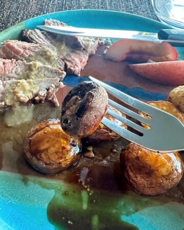 A shiny fork holds up a whole grilled mushroom over a turquoise dinner plate with slices of steak and round baby potatoes.