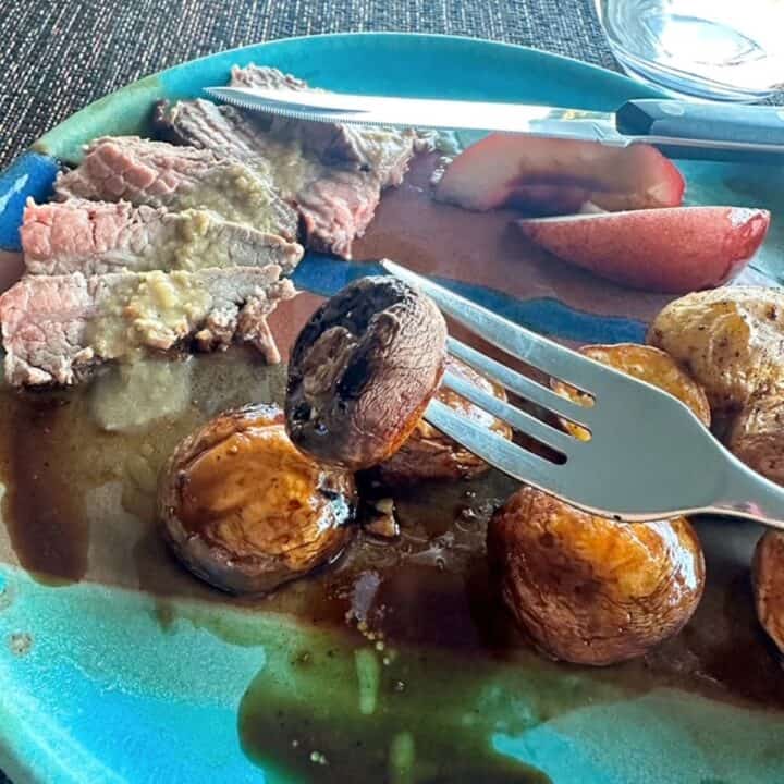 A shiny fork holds up a whole grilled mushroom over a turquoise dinner plate with slices of steak and round baby potatoes.