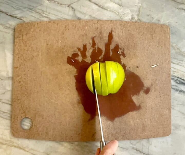 Brown cutting board holds half a green apple being sliced.