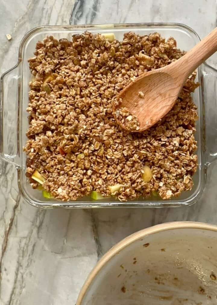 Brownish, clumpy granola topping unbaked over apple filling in clear, square baking dish.