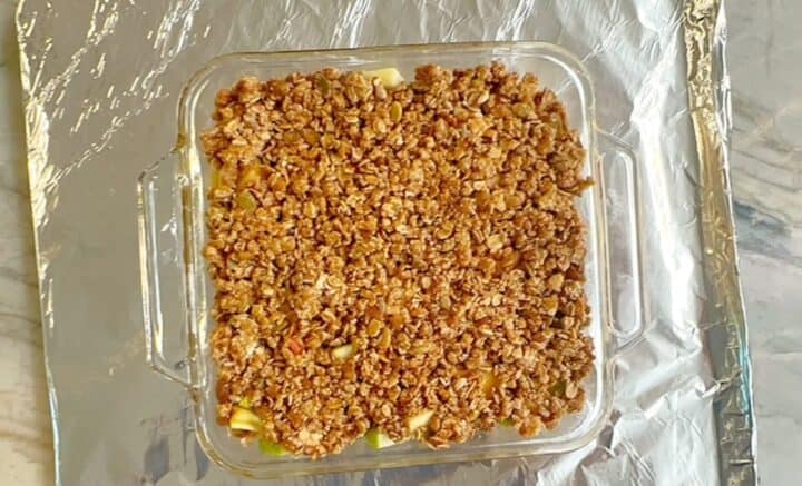 Square clear dish with unbaked apple crisp on top of a foil-lined baking sheet.