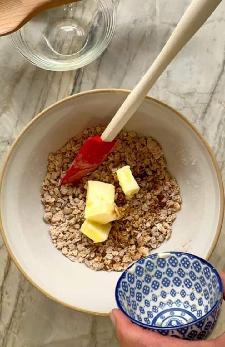 Brownish topping for apple crisp in a white bowl with yellow pats of butter on top.