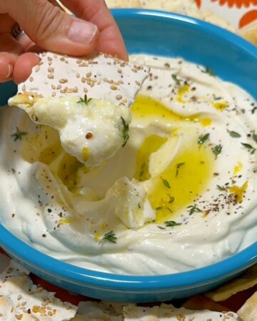 A light sesame seed studded flatbread dips into a creamy ricotta dip in a green bowl.