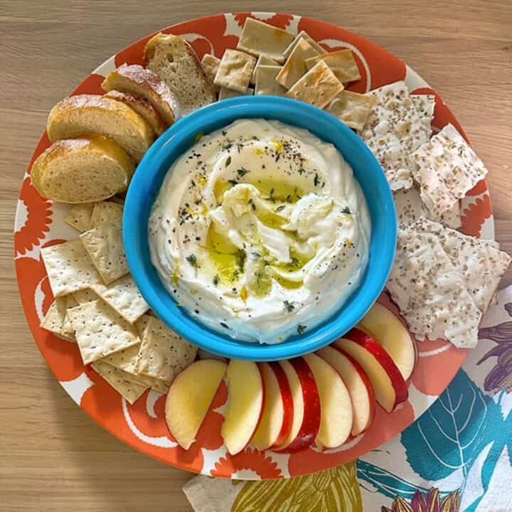 A green bowl full of creamy ricotta dip is surrounded by crackers and apple slices on an orange and white platter.
