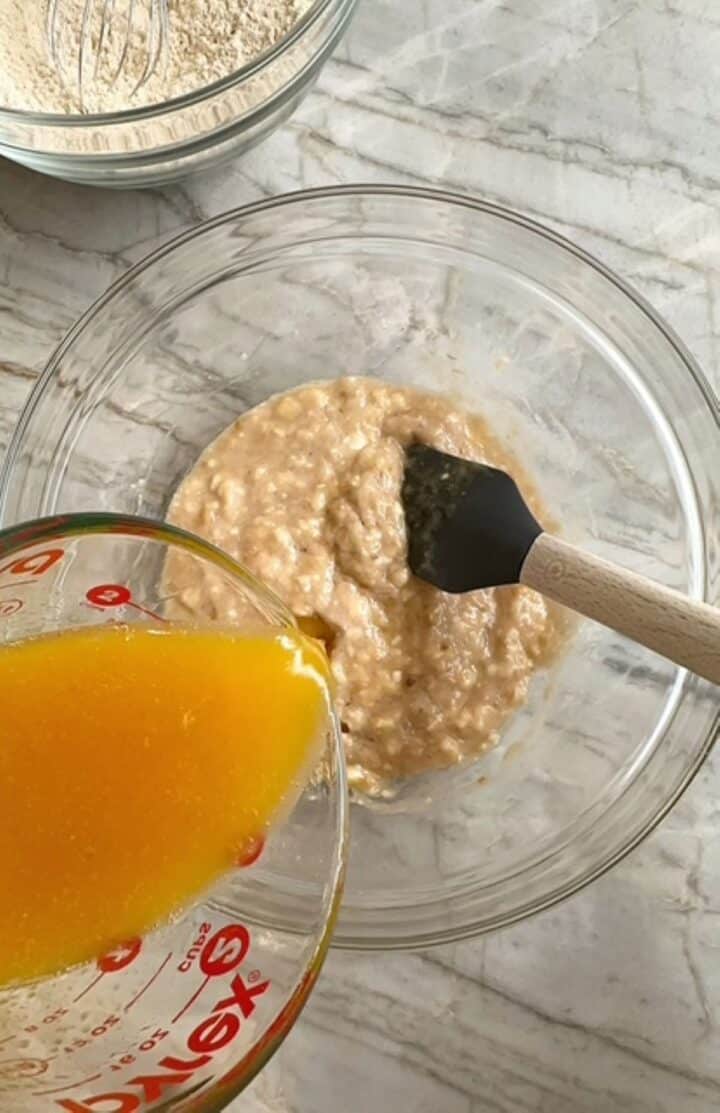 Yellow liquid ingredients are added to mashed bananas in a clear bowl for banana muffins.