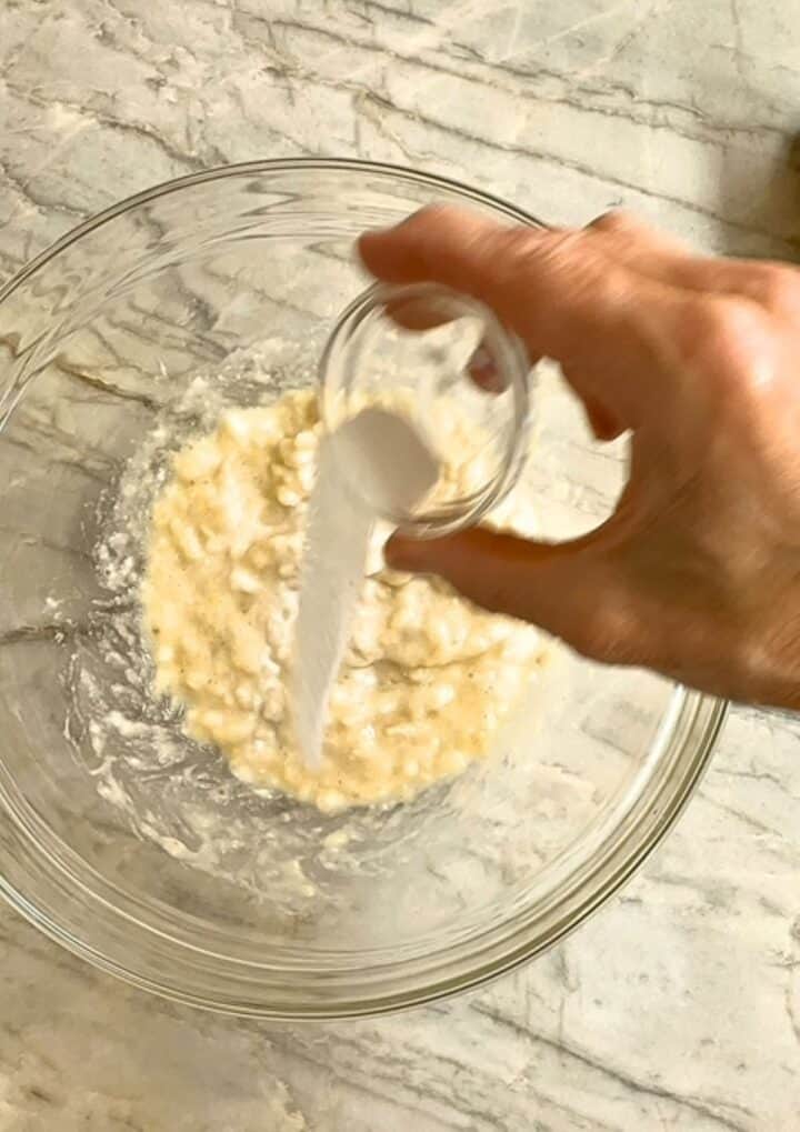 Hand pours white baking soda into bowl of mashed bananas