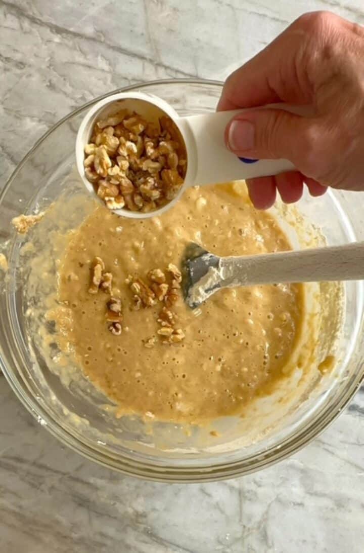 Dumping brown, walnut pieces into yellow banana muffin batter.