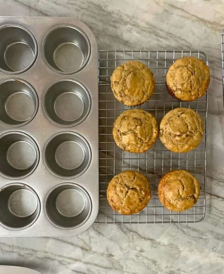 Baked gluten-free golden banana muffins on a wire cooling rack next to gray muffin tin.