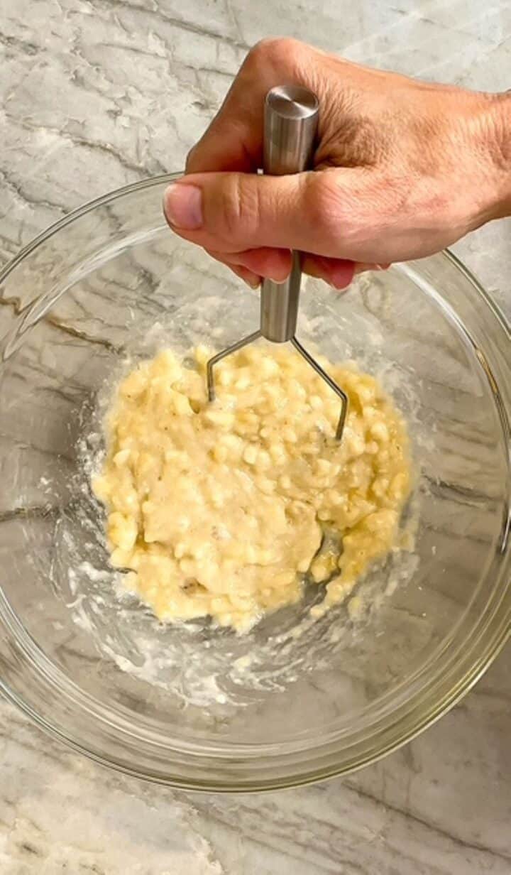 Clear bowl holds bananas being mashed with small silver potato masher.