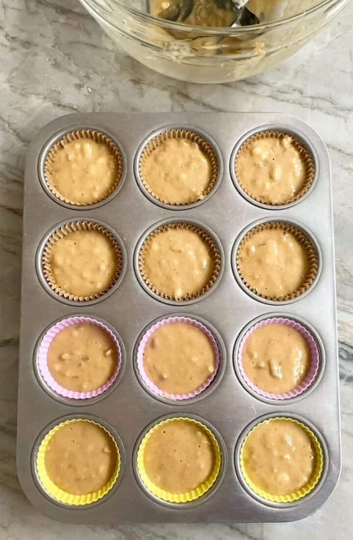 Yellow-tan banana muffin batter fills the liners in a gray muffin tin.