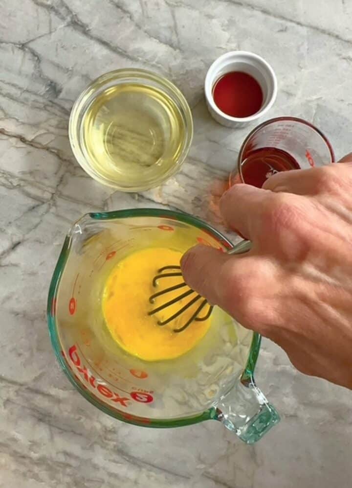 Yellow eggs are beaten by a whisk in a clear liquid measuring cup.