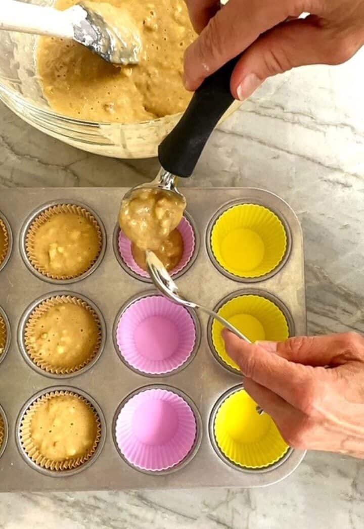 Scooping yellow banana muffin batter into cupcake liners in a gray muffin tin.