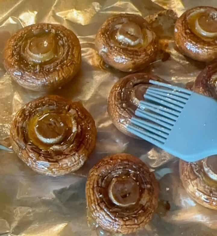 Cooked whole brown mushrooms on shiny foil getting glazed with a blue silicone brush.