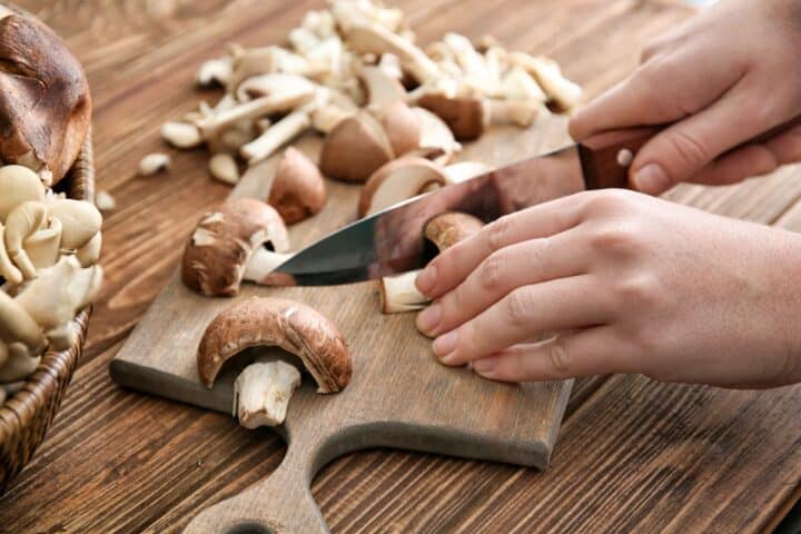 Hand slicing brown muchrooms on a light brown wooden cutting board.
