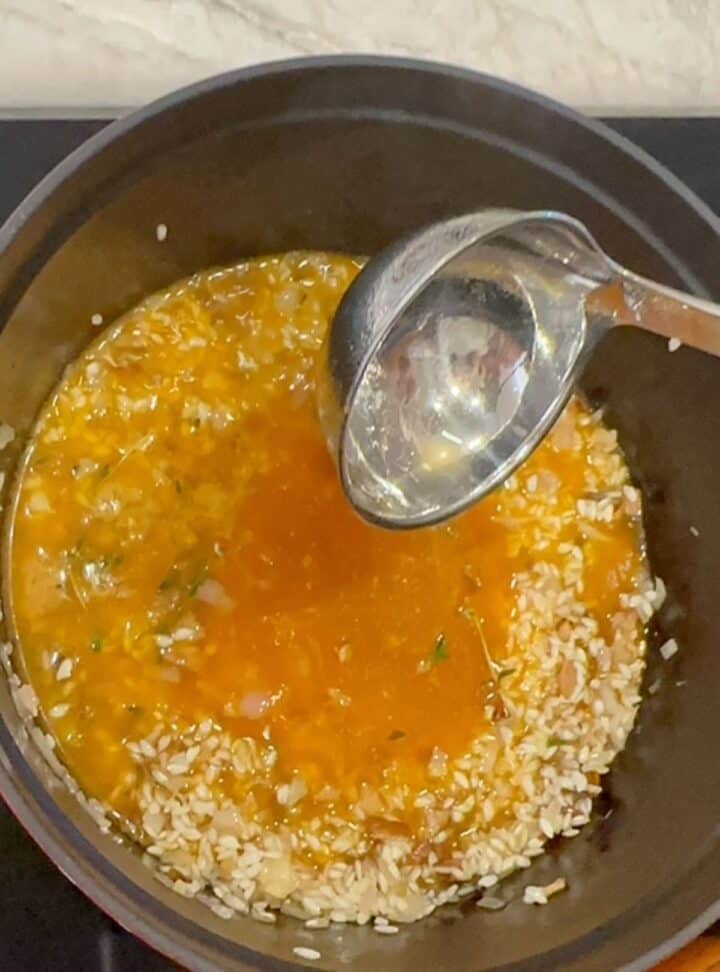A shiny ladle deposits golden veggie broth into light risotto rice mix in a round pot with black interior.