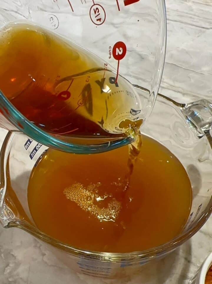 Pouring brown mushroom soaking liquid into a large clear measuring cup of golden vegetable broth.