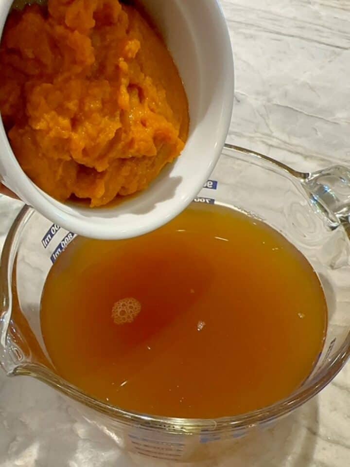 Orange pumpkin puree is added to a large clear measuring cup full of broth.