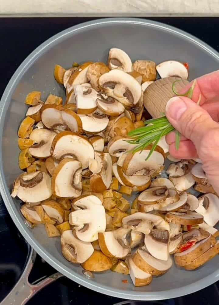 Hand drops fresh, green rosemary into a skillet with sliced brown mushrooms and beige chicken sausage pieces.