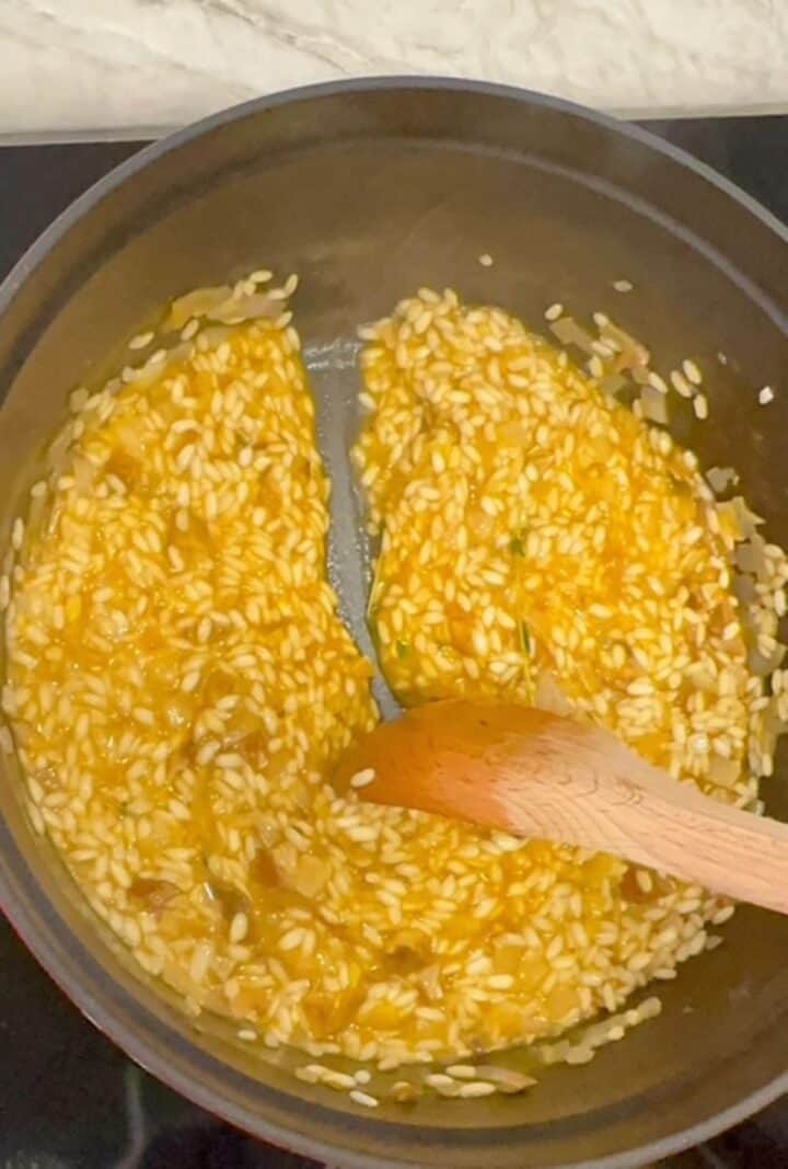A wooden spoon pulls through golden risotto rice, leaving a bare mark on the bottom of a round pot with black interior.