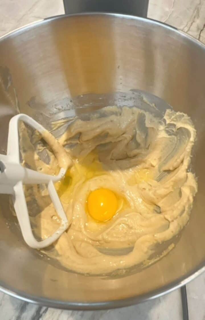 A white paddle beater lays in a metal bowl with a raw egg floating in light, sugary batter.