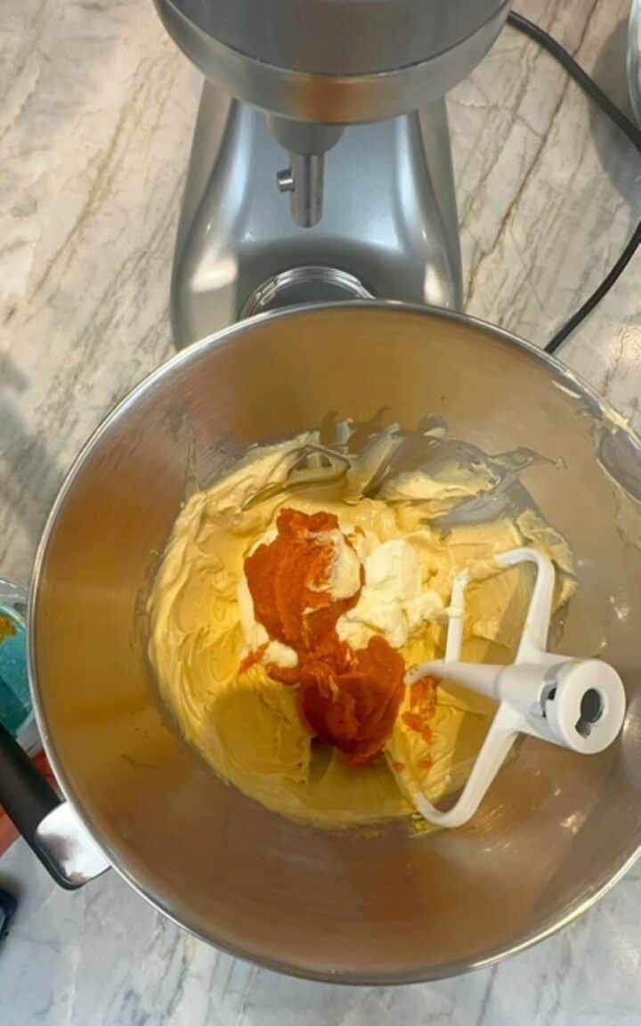 Dark amber pumpkin puree and white ricotta cheese are added to a light colored batter in a stand mixer bowl.