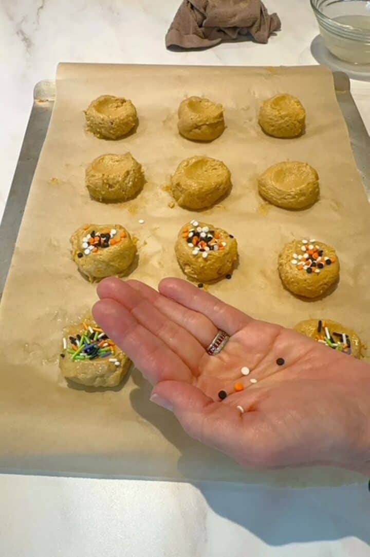 Halloween sprinkles rest in an open palm next to a baking sheet lined with brown parchment and lined with golden pumpkin dough balls.