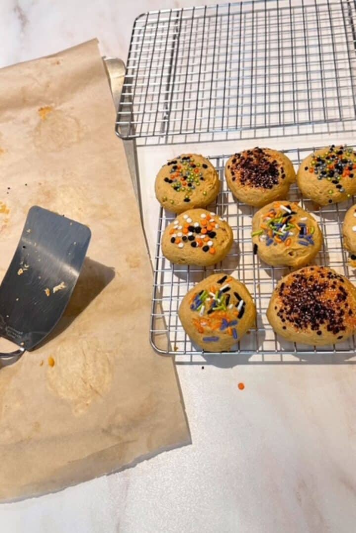 Wire cooling rack holds golden pumpkin ricotta cookies with seasonal sprinkles and a spatula is off to the side.