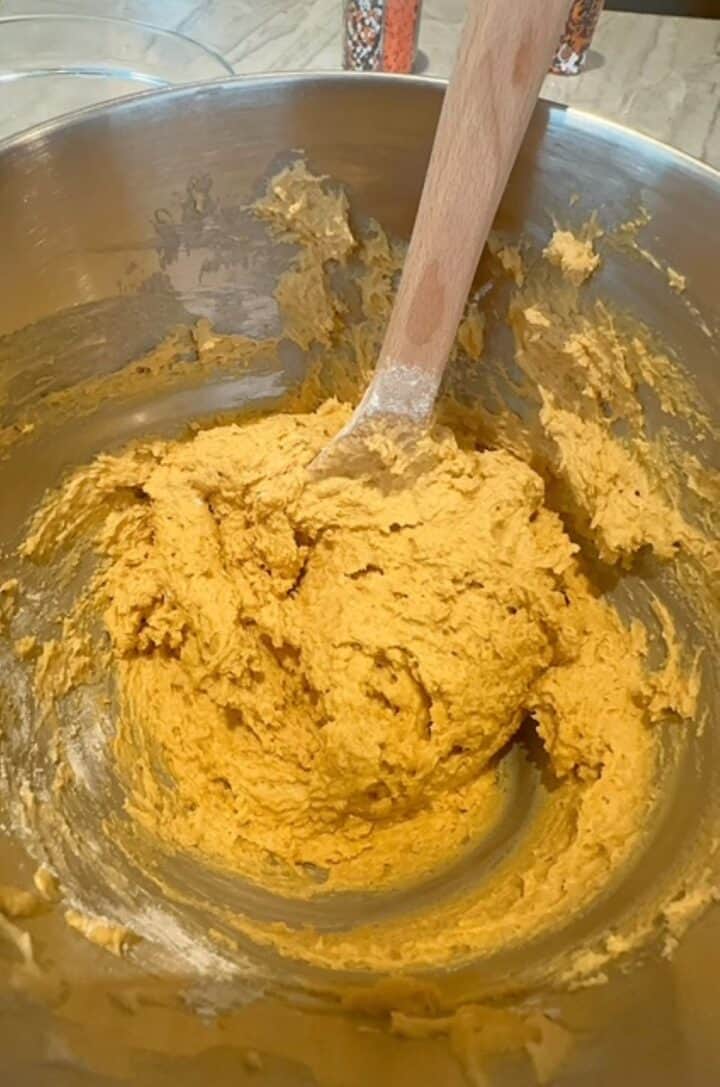 A metal bowl with a brown wooden spoon holds a light orange dough for Pumpkin Ricotta Cookies.