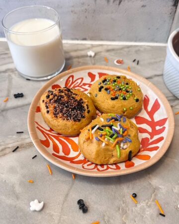 Golden Pumpkin Ricotta Cookies on an orange and white patterned plate are decorated with Halloween sprinkles. A small clear glass of white milk is behind the plate.