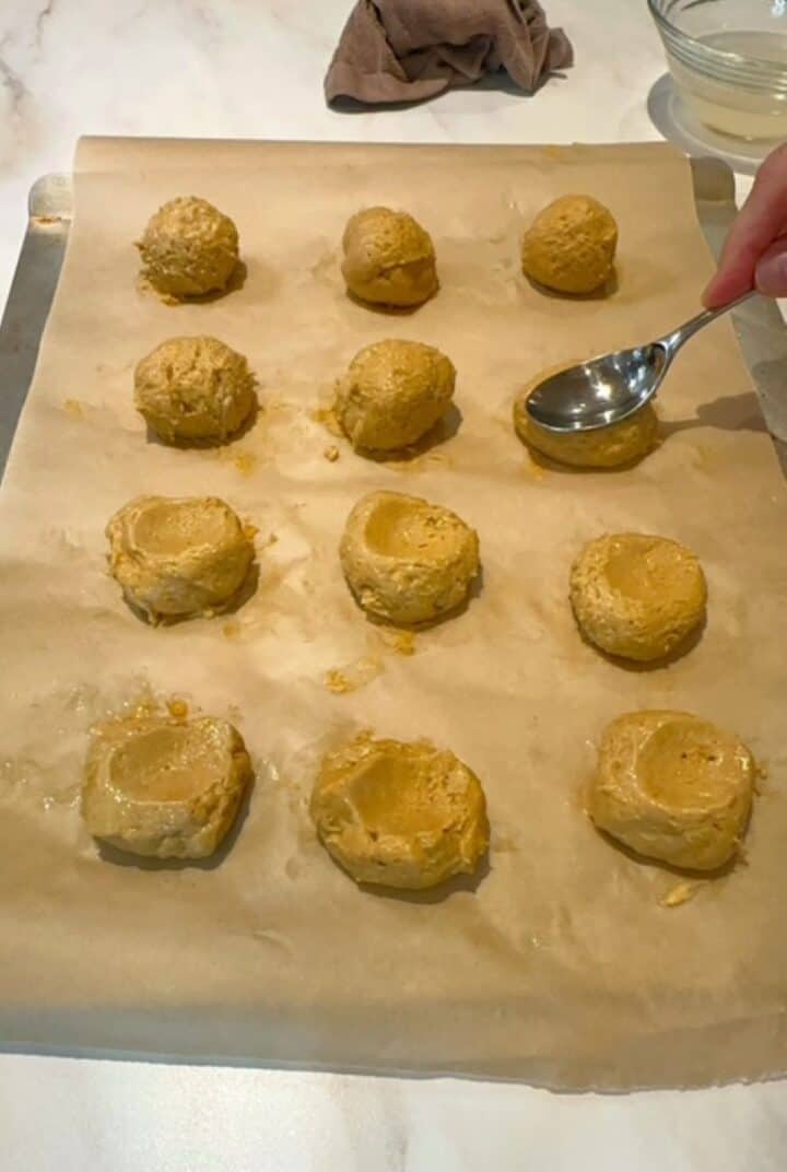 Indentations are made in golden pumpkin balls of cookie dough with the back of a chrome spoon.