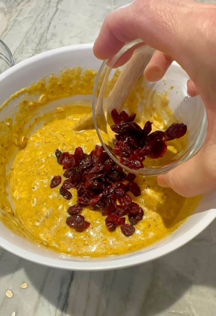 Hand pours red dried cranberries into creamy orange pumpkin oat batter in white bowl.