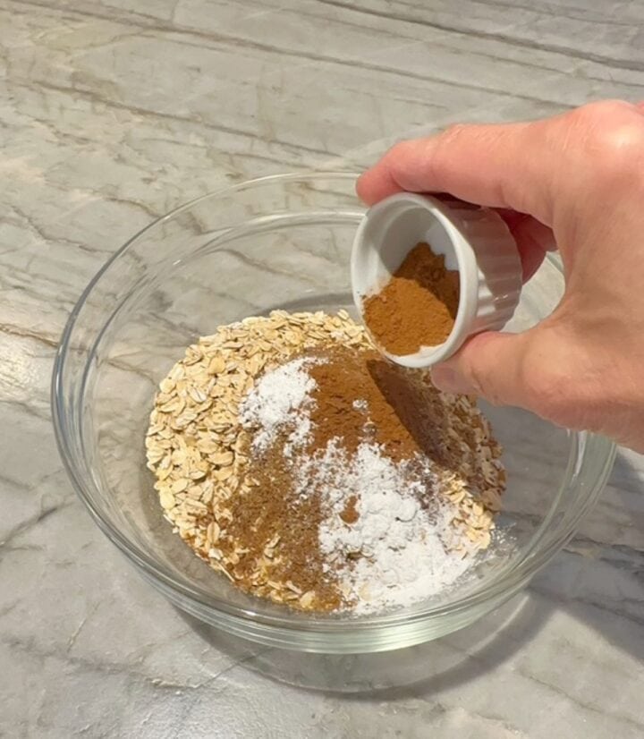 Hand pours brown pumpkin spice over oats, white baking soda, etc in clear bowl,